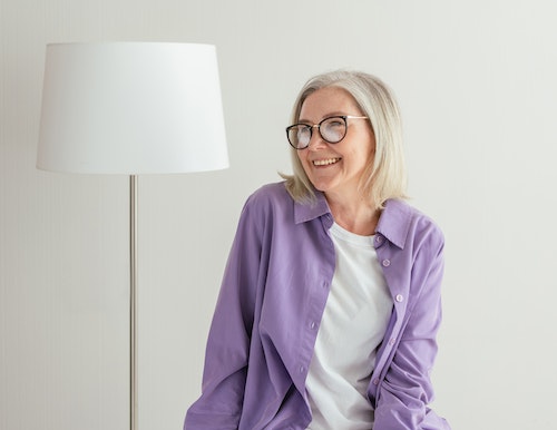Older female wearing white shirt and purple button up looking to the side smiling with lamp in the background - copd home care los angeles
